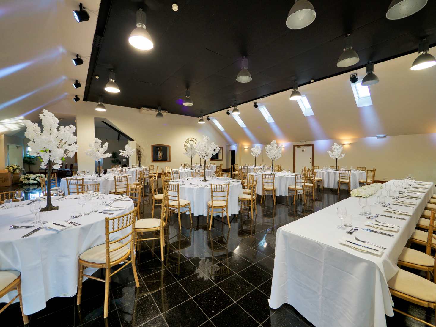 A white wedding table layout on a black floor with gold chairs. 