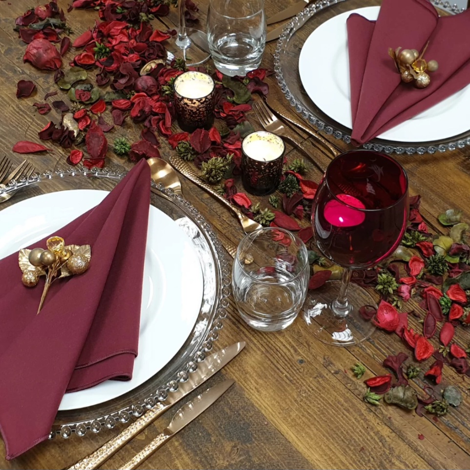 Two Valentine's place settings at a wooden table, with white plates, burgundy napkins topped with gold berries, crystal tumblers, red wine glasses and scattered red rose petals.