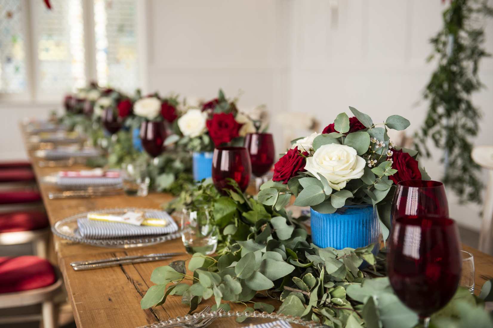A wooden trestle table laid with clear and red wine glasses and blue tin cans filled with red and whire roses and eucalytus.