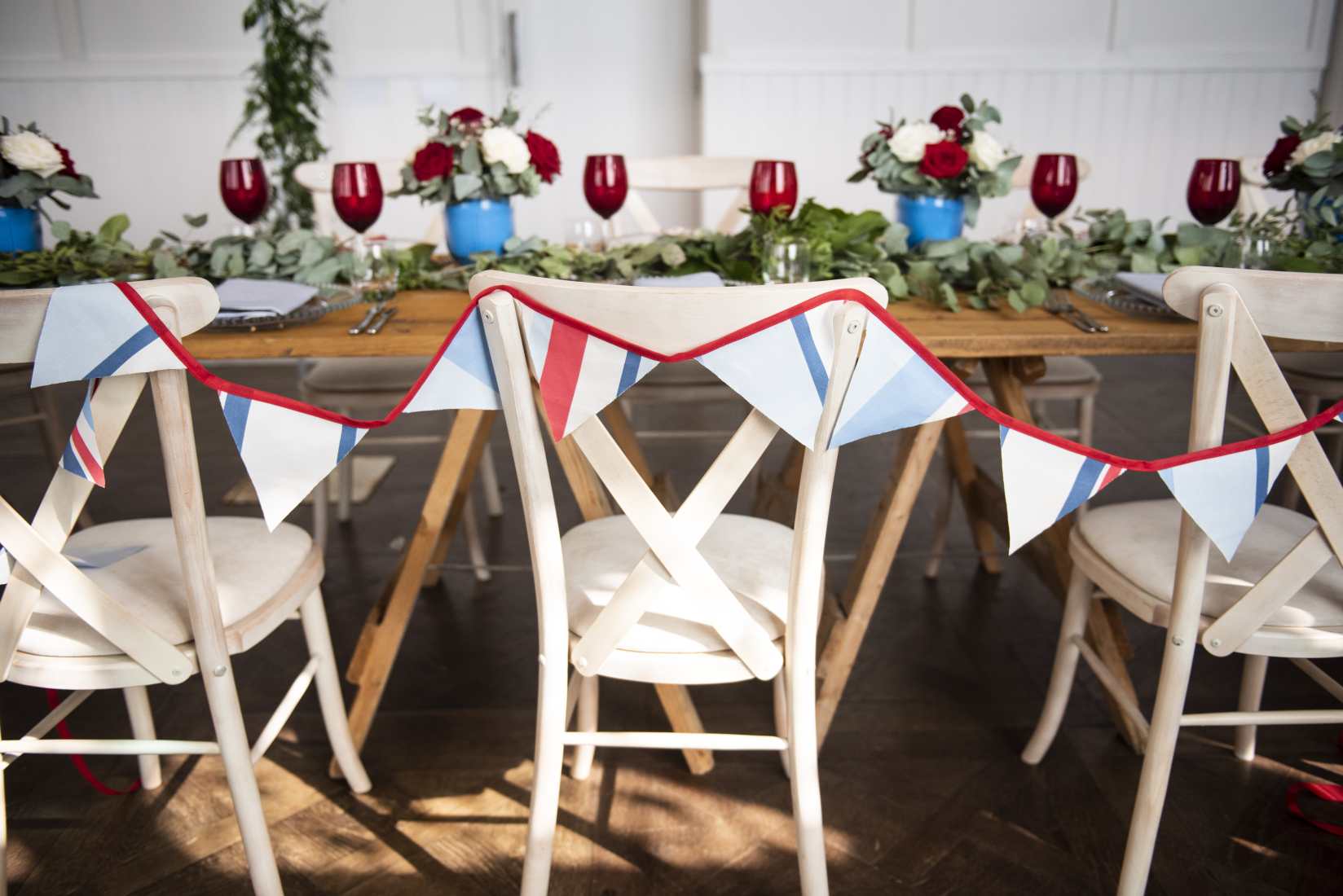 A wooden trestle table laid with mixed red, white and blue crockery, glassware and flowers, plus white chairs decked with bunting.
