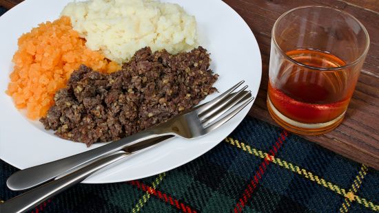 A plate of haggos, neeps and tatties, and a glass of whisky, on a blue tartan place setting.