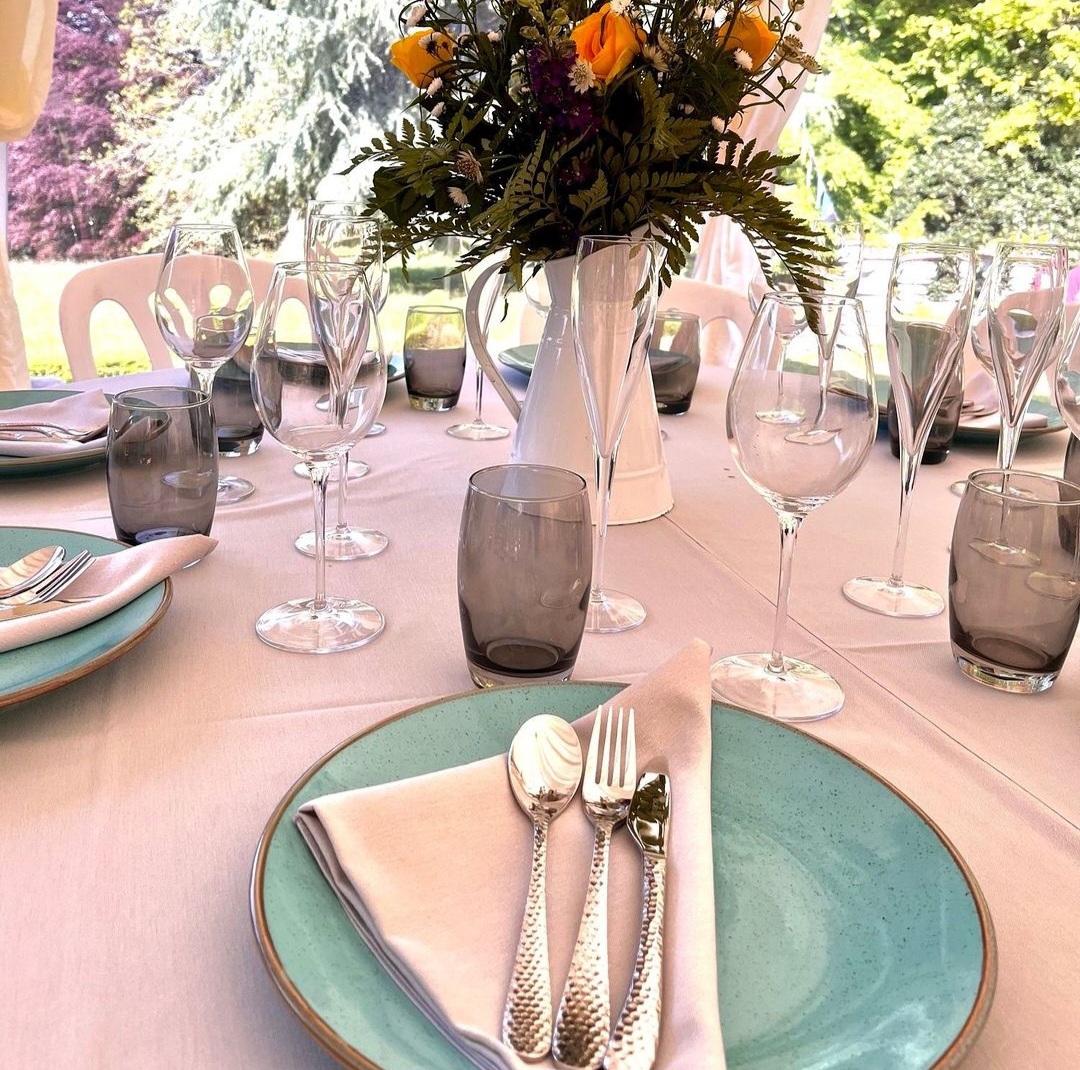 A garden wedding table laid with aquamarine plates, rose gold cutlery, smoke tumblers, and striking orange flowers.
