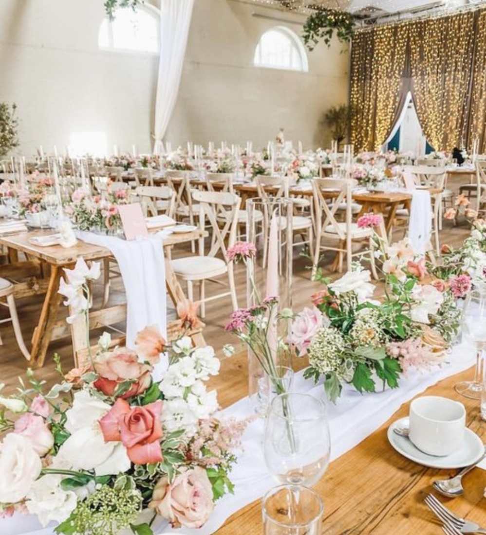 A suite laid for a wedding breakfast, with light wood tables, white chairs, crystalware, silver cutlery and beautiful pink blooms.