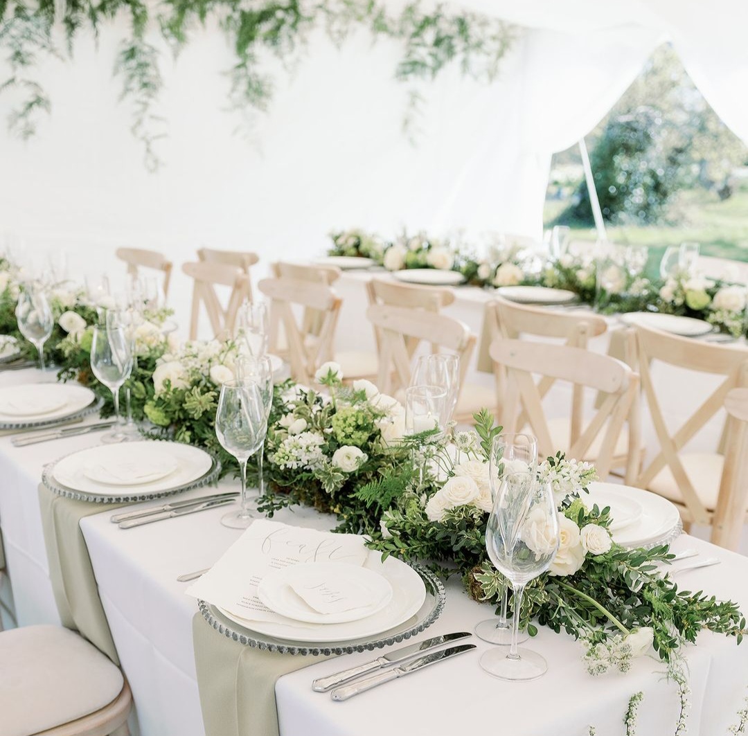 Trestle tables in a white marquee for a wedding, with white table linen, crockery, crystalware, and green foliage.