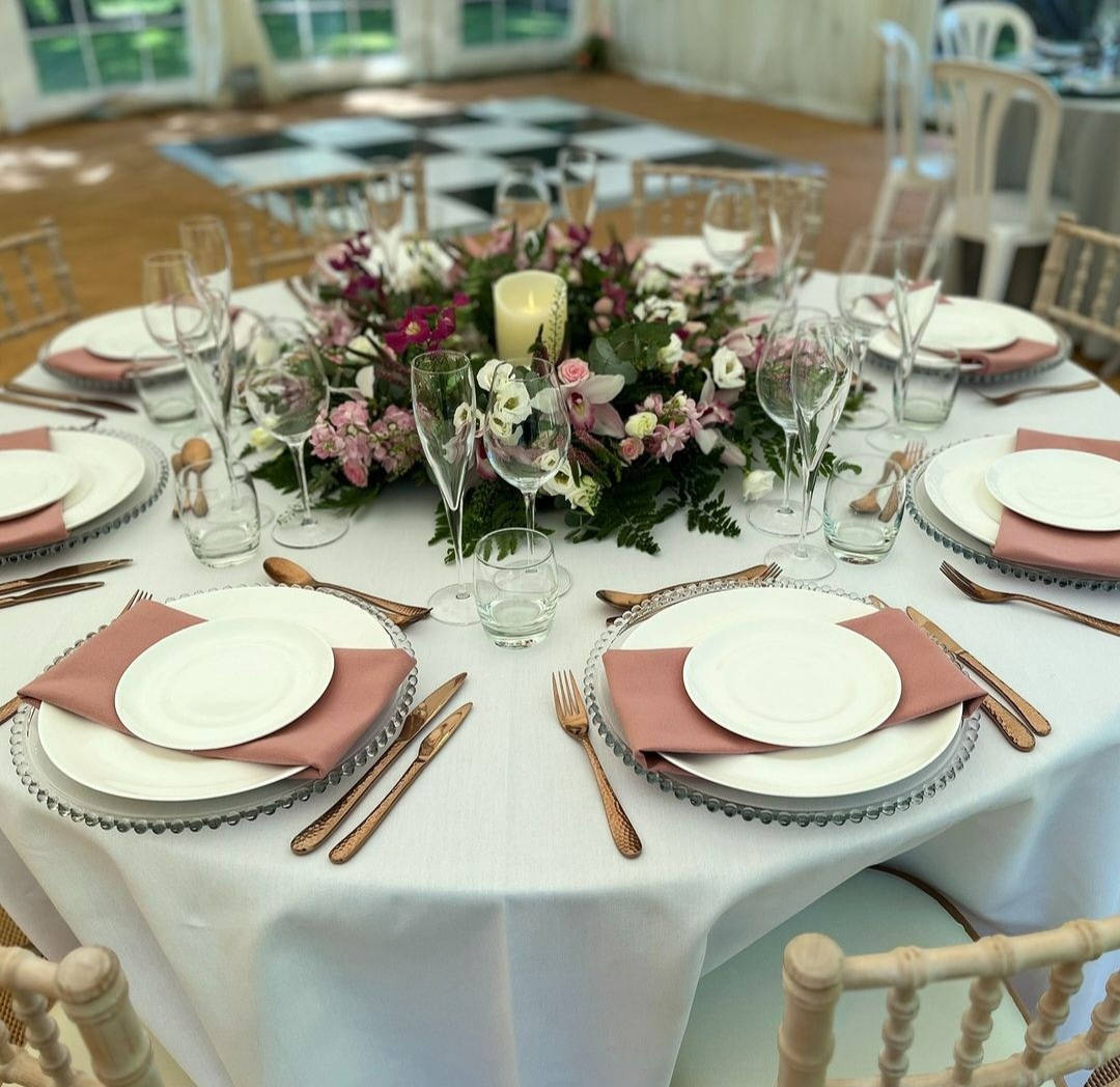 Wedding table with white chairs and cloths, disky pink napkins, rose gold cutlery, and a burgundy themed floral centrepiece.