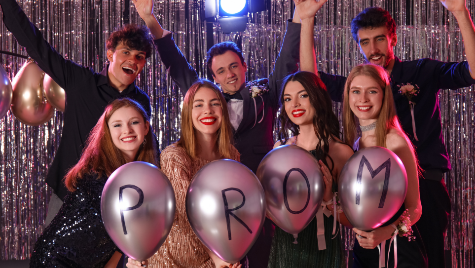A group of smiling students holding up silver balloons spelling out PROM.