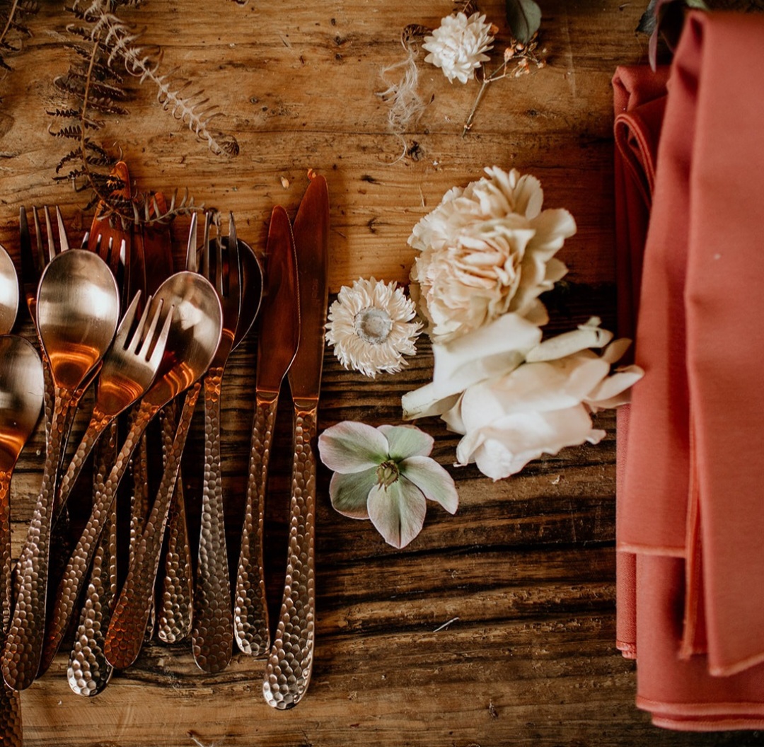 A pile of Liza Rose cutlery and peach napkings on a rustic table.