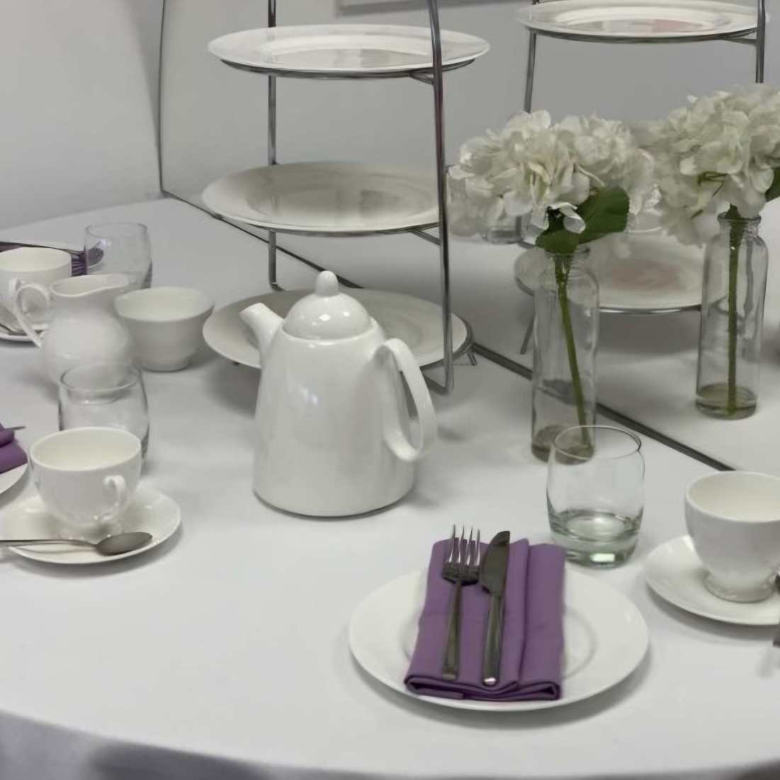 Classic afternoon tea crockery including a cake stand, reapot and simple whire flowers in small glass vases, on a white tablecloth with a lilac napkin and silver cutlery.