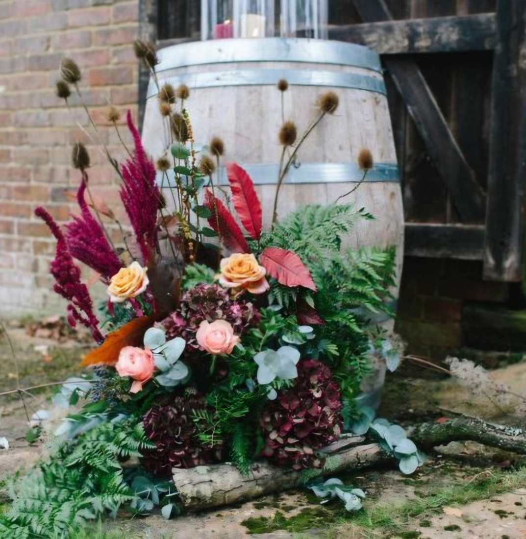 A wooden barrel with a spectacular burgundy, peach and sage green floral arrangement in front of it.