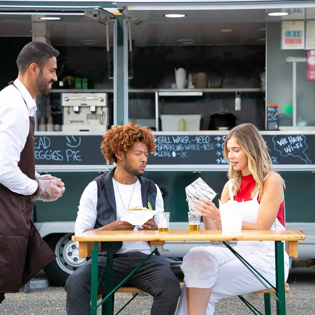 A young mixed race couple enjoying street food and drinks, sat in front of a food truck.
