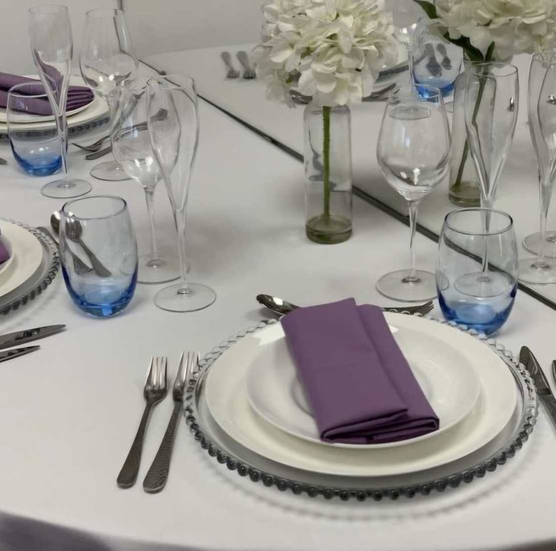 A table set with a white table cloth, white flowers, China crockery, crystal glassware and a lilac napkin for a Mother's Day brunch.
