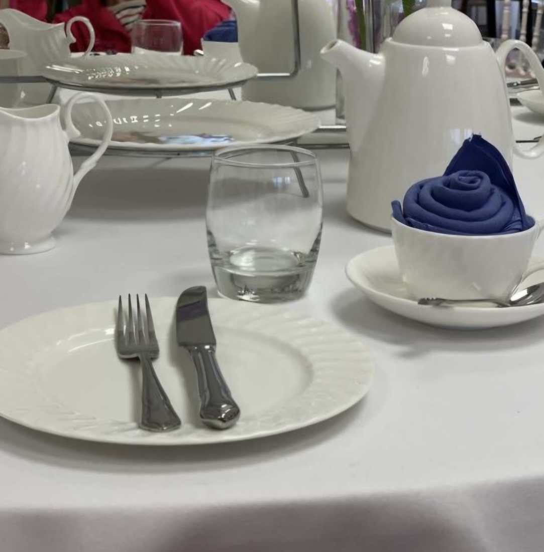 A table set with a white table cloth, white China crockery, glassware, silver cutlery and a nany blue napkin wrapped like a rose for a Mother's Day brunch.