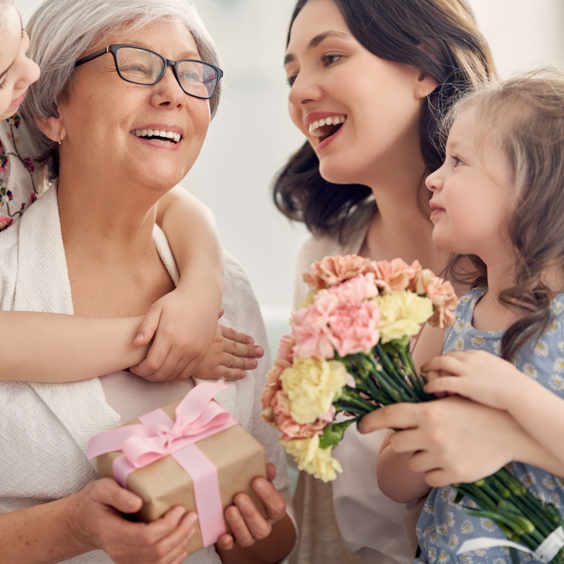 A grandmother smiling, with her daughter and two small granddaughters giving her a ribbon-wrapped present and a bunch of carnations for Mother's Day.