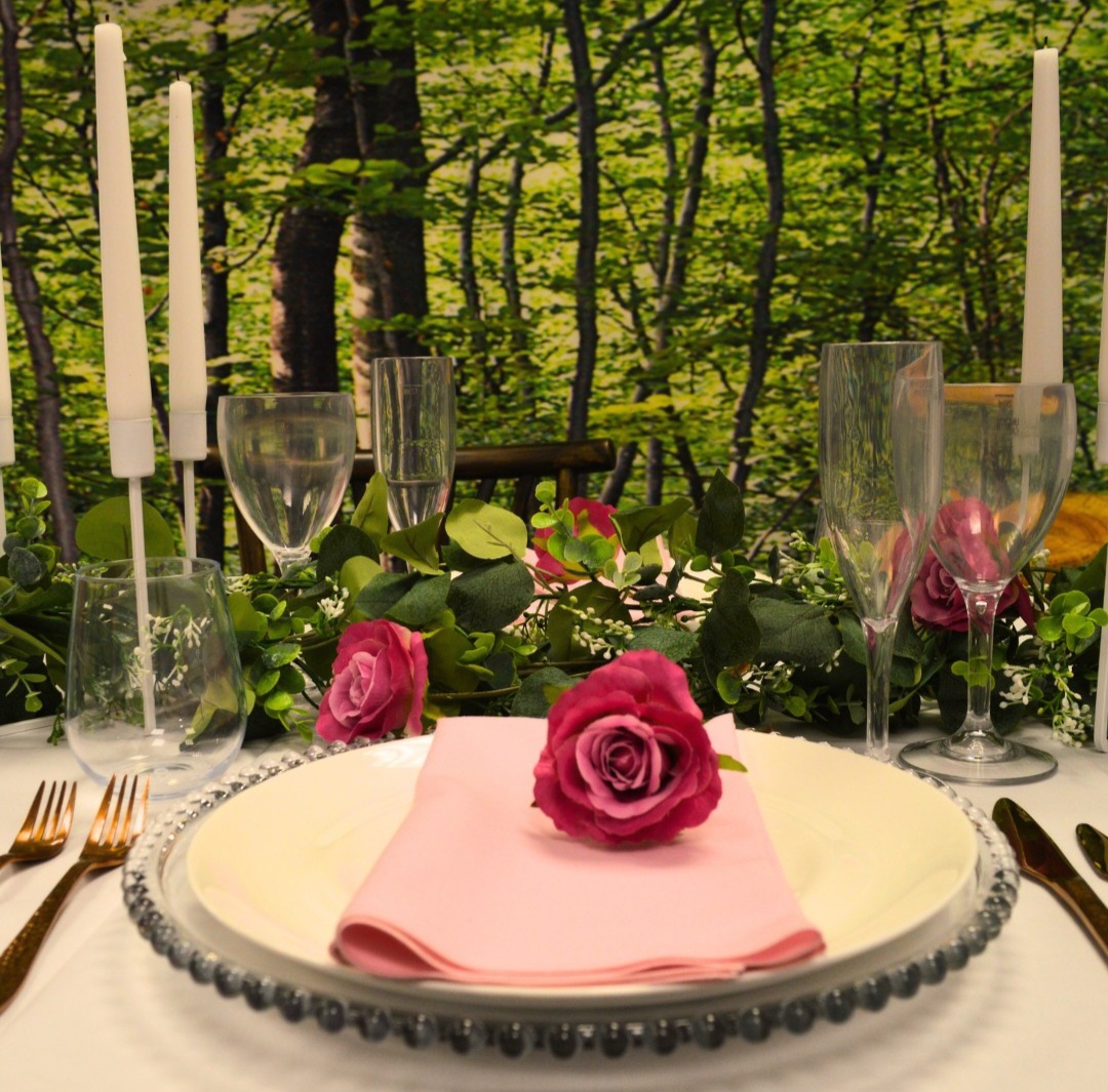 A springtime table setting outdoors in a woodland setting, with a white plate on a clear glass charger, topped with a light pink napkin and dusky pink rose, wih Liza rose gold cutlery, crystal champagne flutes and wine glasses, and white taper candles.