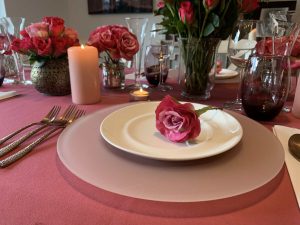 A place setting at a table for a Valentine's dinner at home, with fancy crockery and cutlery, candles and lots of red roses.