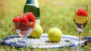 A tennis racket, tennis balls, bottle and glasses of bubbly and a bowl of strawberries on a tennis court.