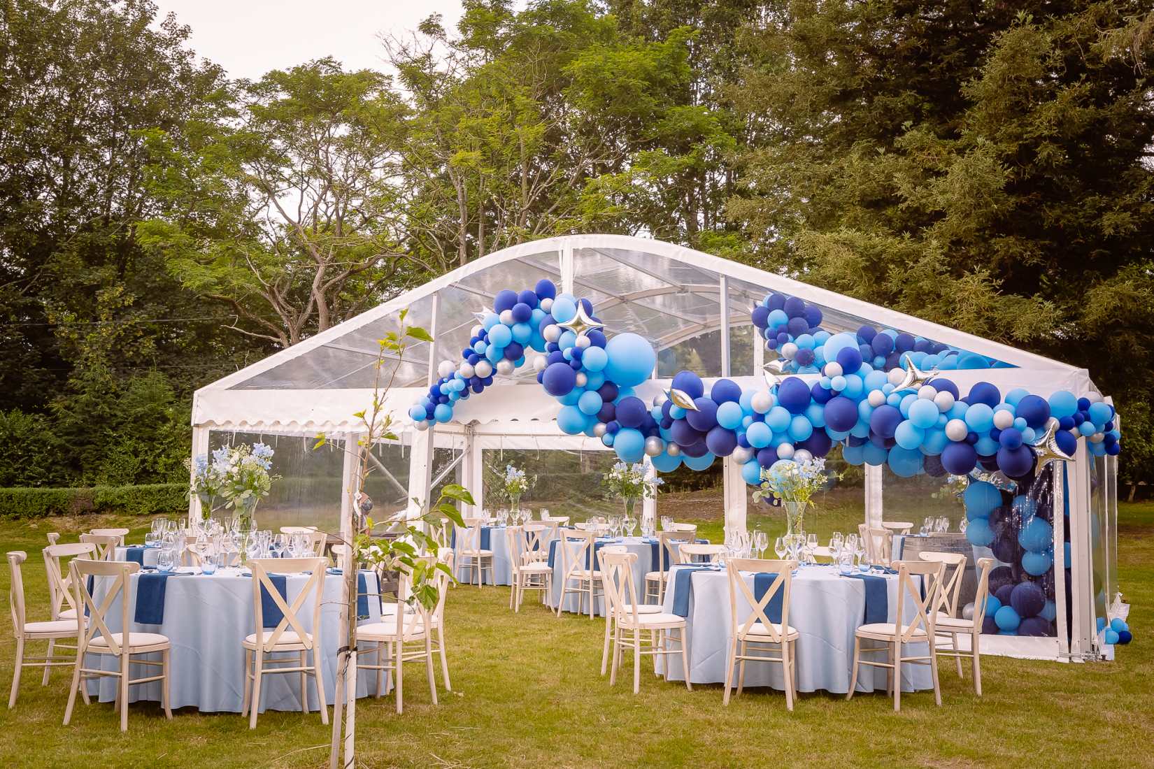 An outdoor transparent marquee with tables and chairs inside and out, bedecked with light and dark blue balloon decor.