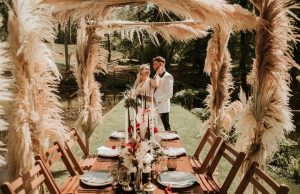 A fashionable bride and groom next to a rustic wooden wedding table and chairs, underneath a canope of giant cream plush feathers.