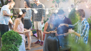 A group of adults in summer clothes eating and drinks under festoon lighting at a company summer party.