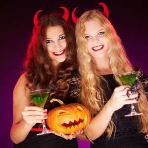 Two glamourous young women in black evenig dressing and devils horns, holding green cocntails and pumpkins.