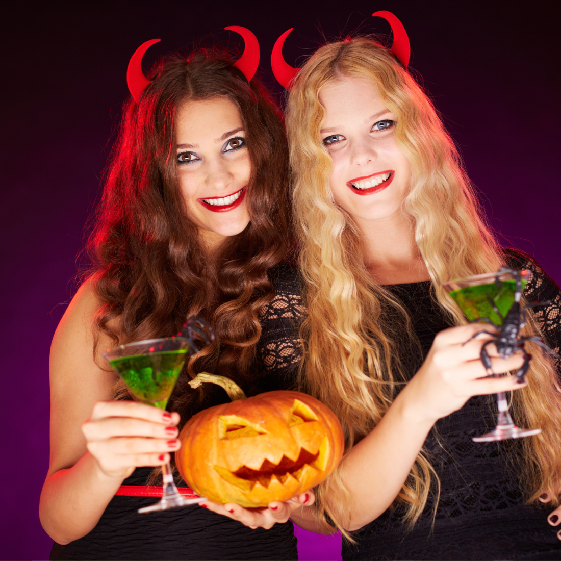 Two glamourous young women in black evenig dressing and devils horns, holding green cocntails and pumpkins.