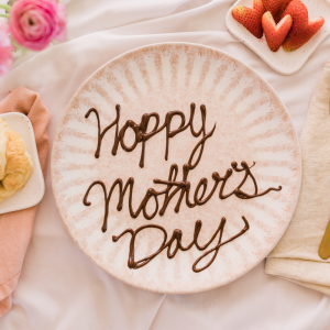 A white plate with Happy Mother's Day piped in chocolate, plus pink flowers and dish of strawberries.