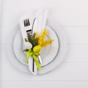 A white plate on a white charger plate, topped with warpped silver cutlery in a white napkin, tied with floral sprigs and mini eggs.