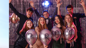 A mixed group of dressed up teenagers at a school leavers prom, holding up four gold balloons spelling out P-R-O-M.